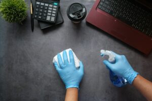 Person in blue gloves cleaning a surface with a spray bottle and cloth, surrounded by a calculator, coffee cup, and laptop, representing professional cleaning services in a post-construction context.