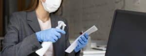 Woman in a mask and gloves cleaning office workspace with spray bottle and documents, emphasizing hygiene for hybrid NYC office environments.